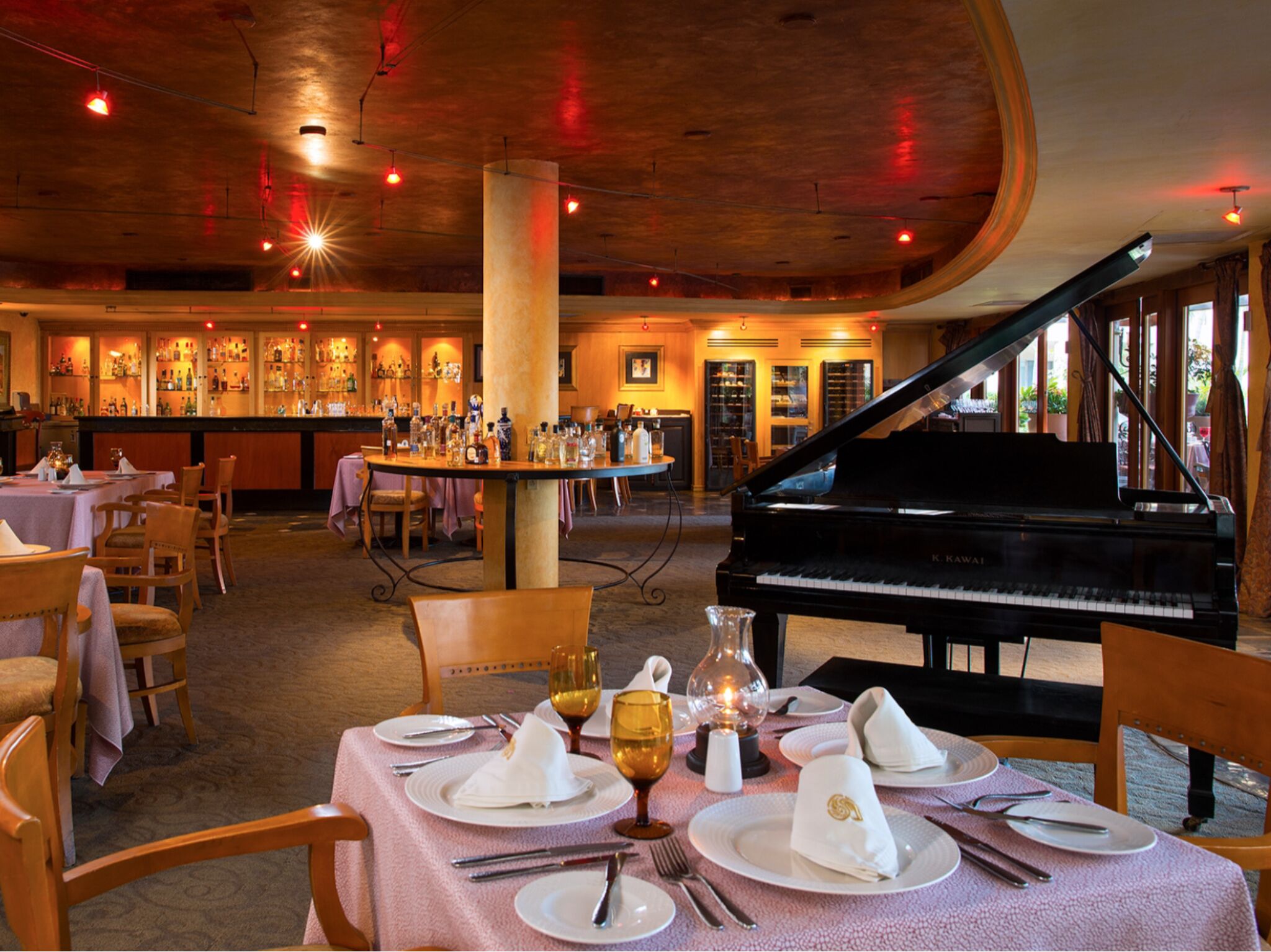 A dining area with a grand piano, tables set for a meal, and a bar in the background.