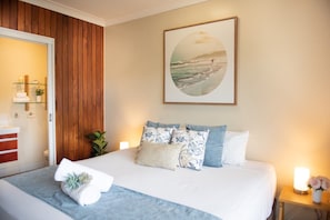 A bedroom with a bed, pillows, a bedside table with a lamp, a wall-mounted picture, and a wooden panel wall. - Eagles Nest On Airlie (Airlie Beach)