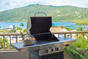 A portable grill with a basket of bread and bottles on a balcony overlooking a lake and hills.