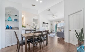 A spacious living area with a dining table, chairs, and a sofa. There is a kitchen area with a countertop and cabinets. A ceiling fan is visible, and the room has a large window with curtains.