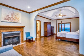A bedroom with a fireplace, a bed, a ceiling fan, and a view through a window.
