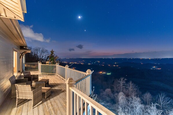 Une terrasse en bois, meublée de mobilier en osier, avec une balustrade d’où l’on peut admirer la vue sur la ville illuminée la nuit.