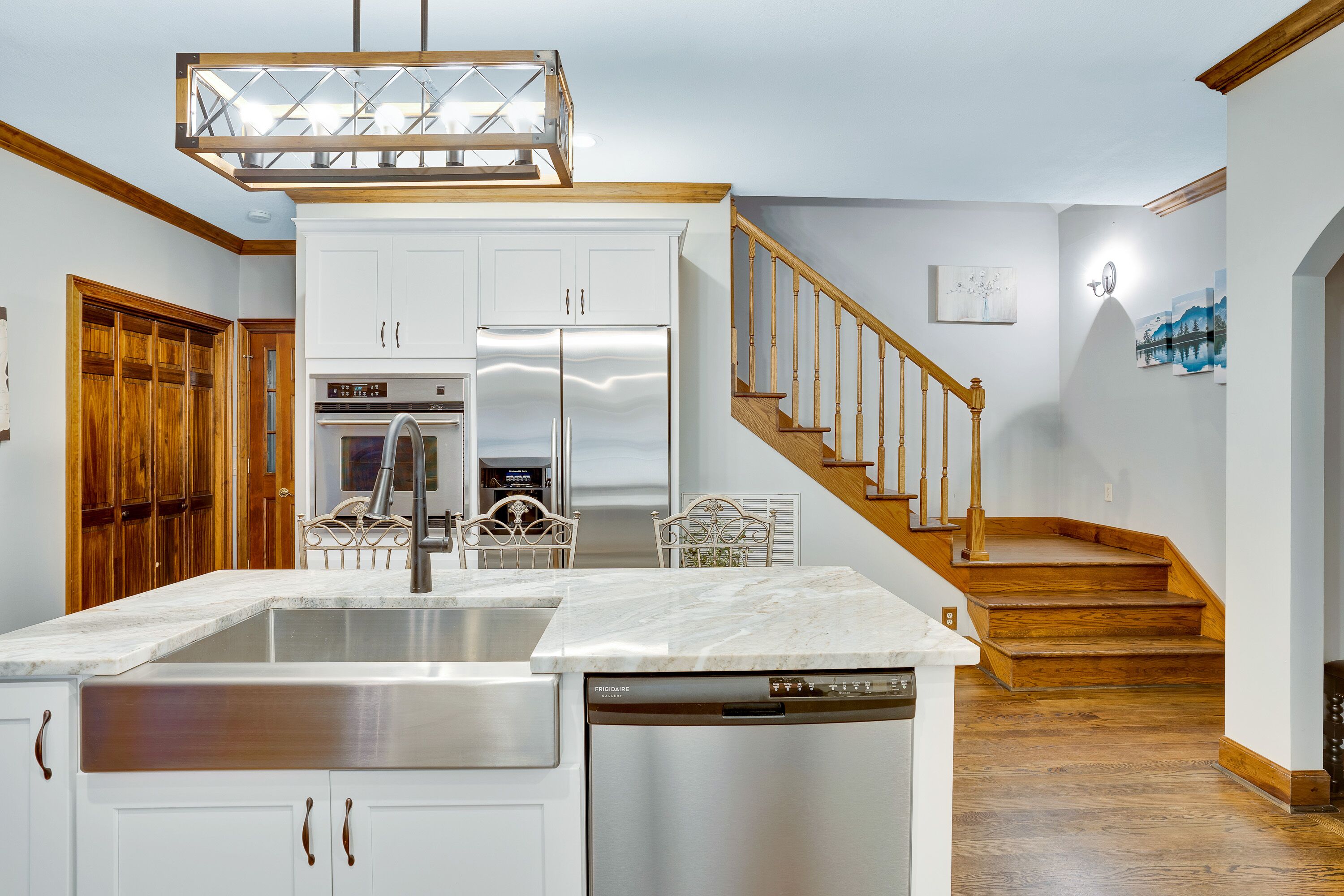 A kitchen with a central island, stainless steel appliances, and a wooden staircase leading to another level.