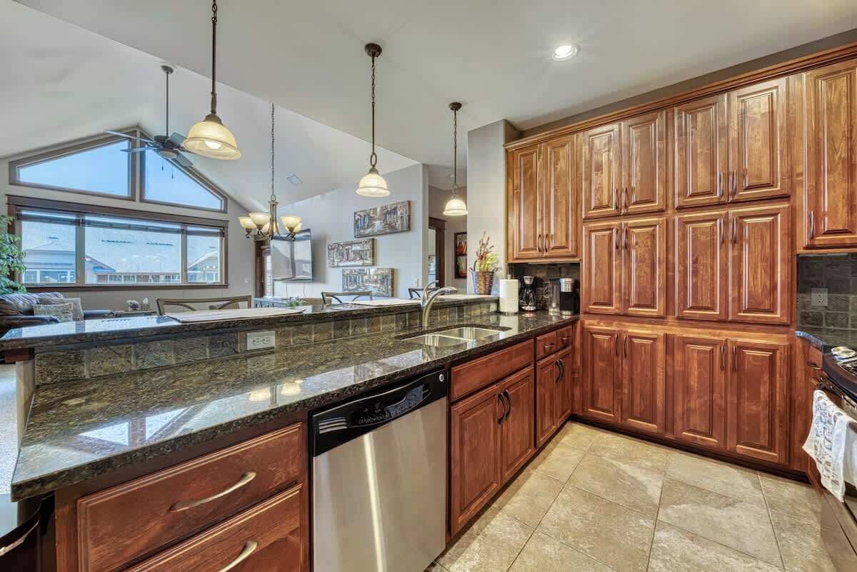 A kitchen with granite countertops, wooden cabinets, stainless steel appliances, pendant lighting, and a large window providing natural light.