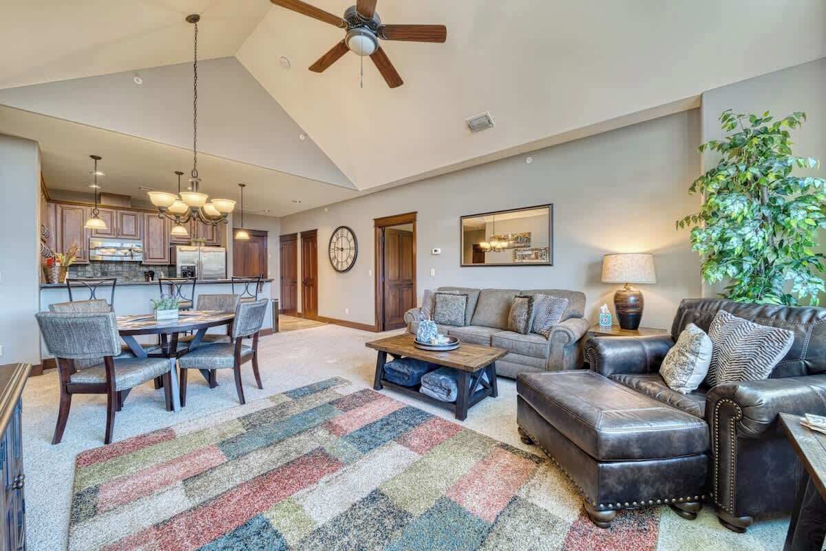 A spacious living area with a large sofa, a coffee table, and a dining table set for six. There is a ceiling fan, a chandelier, and a large plant in the corner.