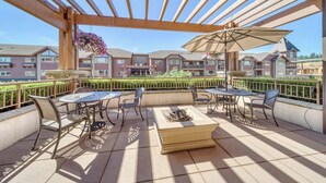 Une terrasse extérieure couverte, avec une table et des chaises, une jardinière suspendue et une vue sur des immeubles d’habitation en arrière-plan.
