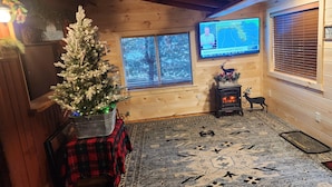 Interior acogedor de una cabaña con un árbol de Navidad decorado, una chimenea y un televisor que muestra un pronóstico del tiempo.