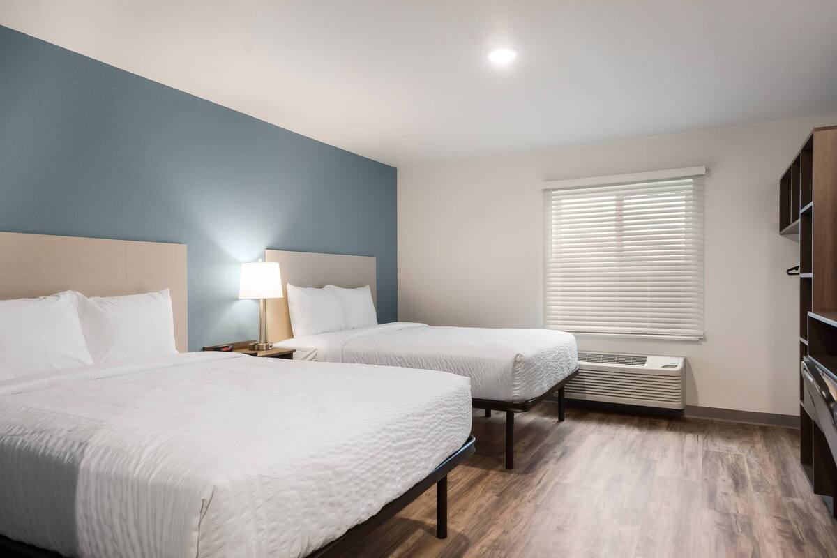A hotel room with two beds, a nightstand with a lamp, a window with blinds, and a built-in air conditioning unit.