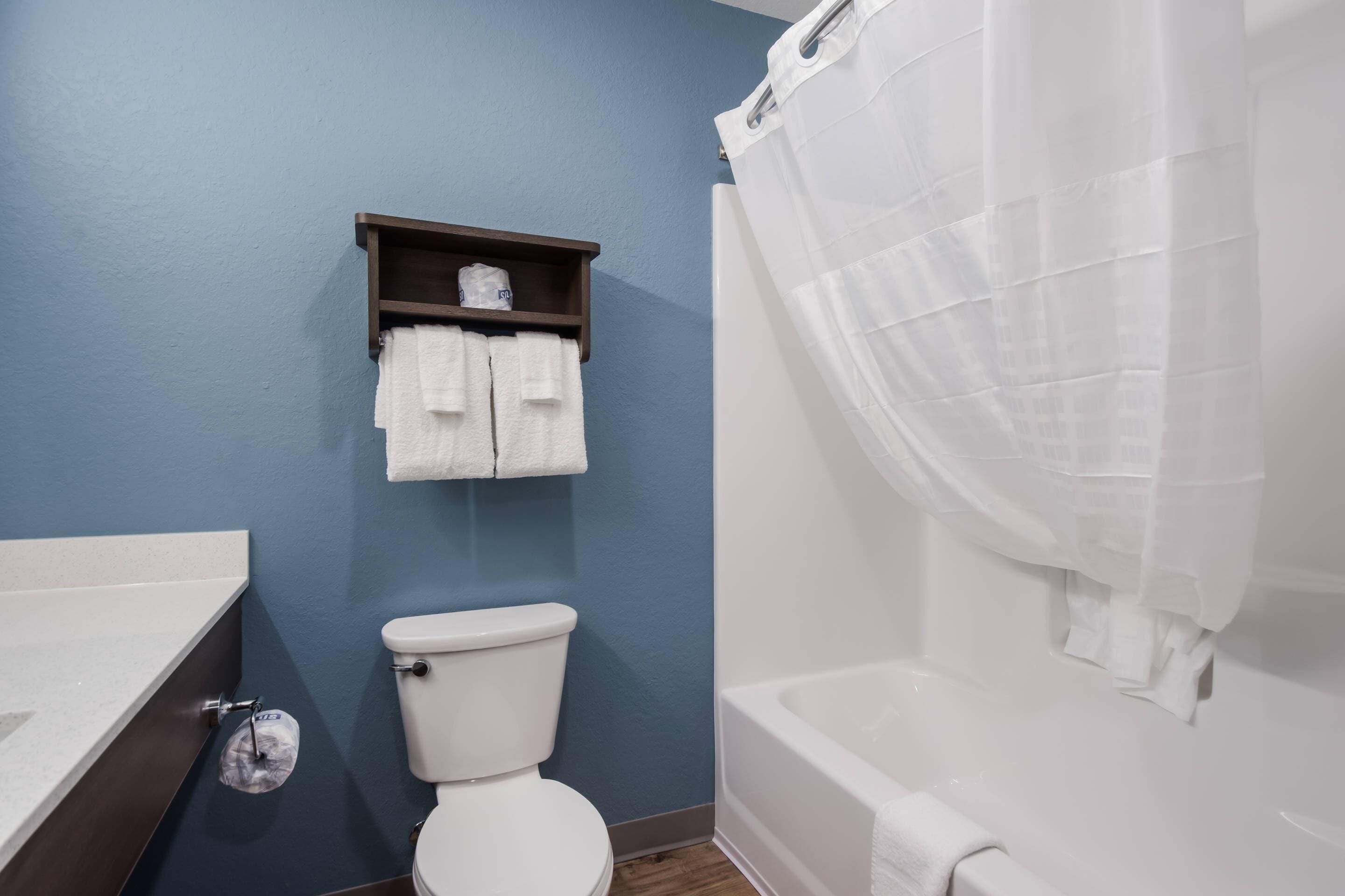 A bathroom with a toilet, a sink, and a bathtub. There is a towel rack with two towels and a shelf with a tissue box.