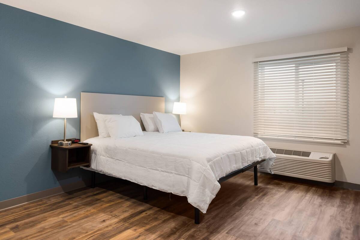 A bedroom with a bed, two bedside lamps, a nightstand, a window with blinds, and a wall heater.