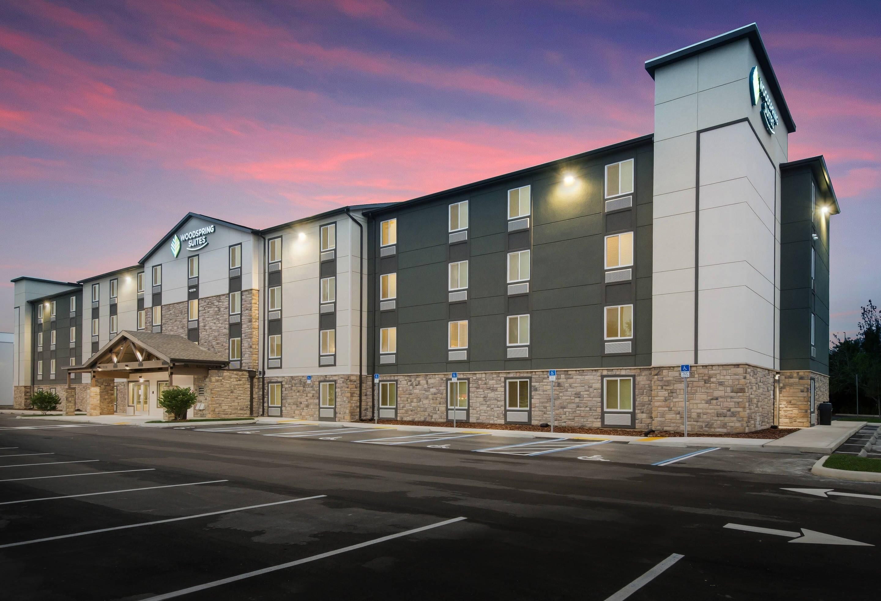 Un hôtel moderne doté d’un toit plat, de nombreuses fenêtres et d’une enseigne indiquant qu’il s’agit d’un Woodspring Suites.