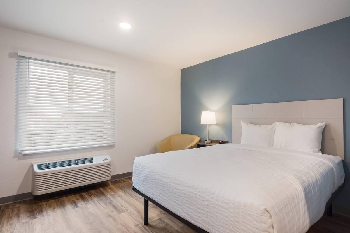 A hotel room with a bed, a chair, a lamp, and a window with blinds.
