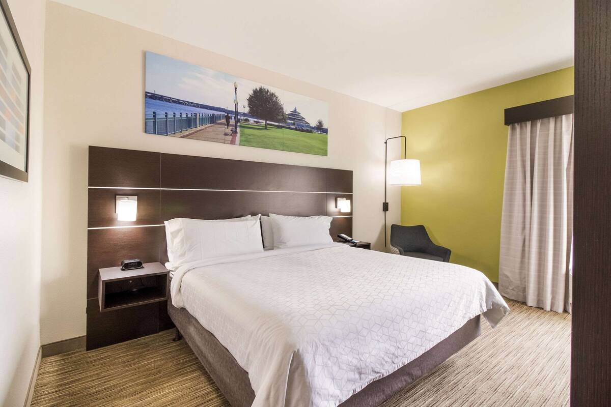 Quarto de hotel com uma cama grande, uma mesa de cabeceira com abajur, uma cadeira e um quadro na parede.