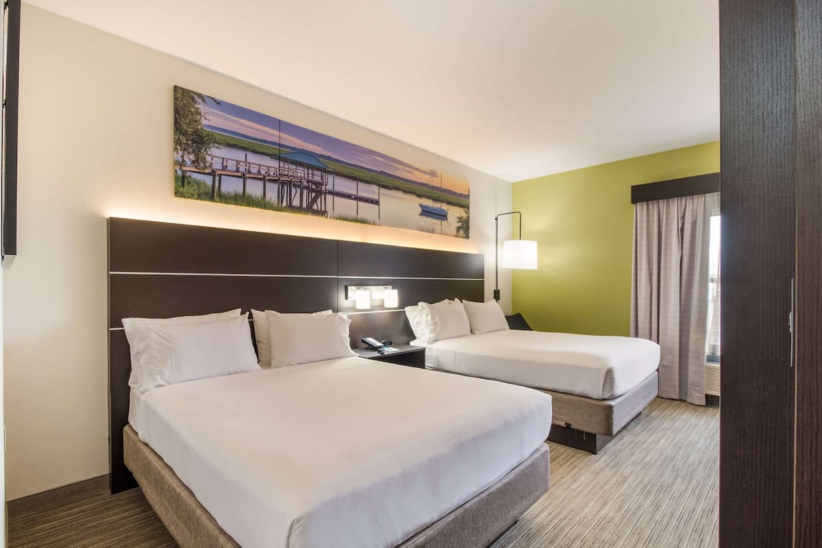 Quarto de hotel com duas camas, uma mesinha de cabeceira, uma luminária e uma pintura de uma paisagem com lago.