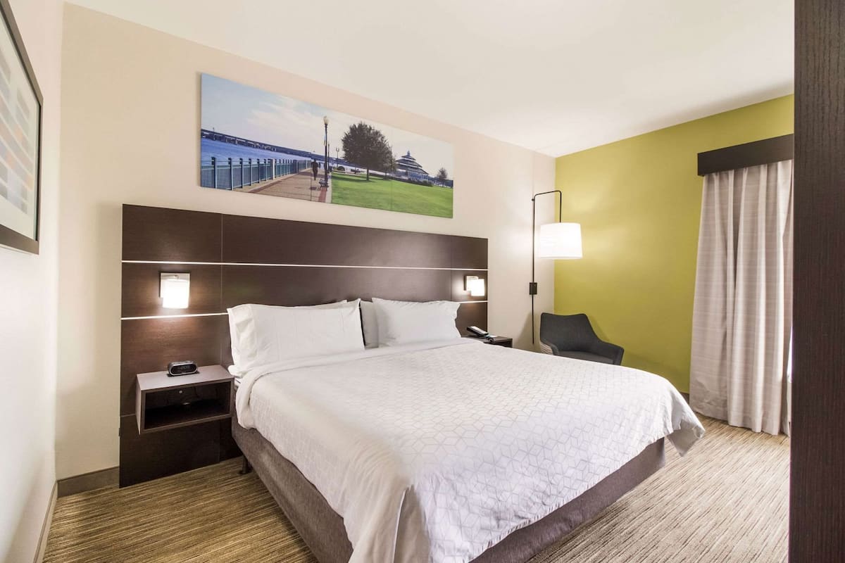 Quarto de hotel com uma cama grande, mesa de cabeceira, abajur, cadeira e um quadro na parede.