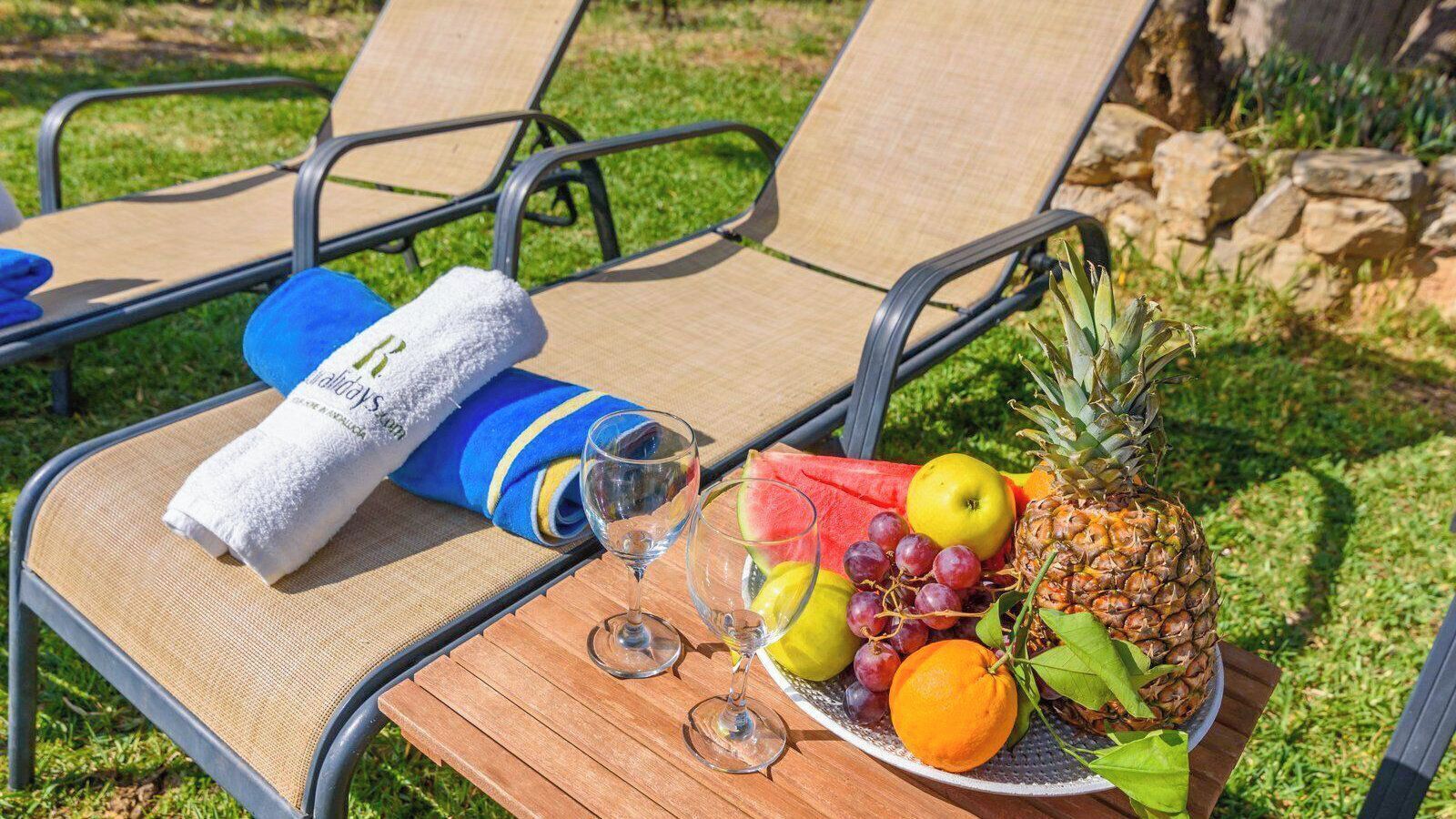 A table with a fruit platter, two glasses, a towel, and a rolled-up towel on a chair.