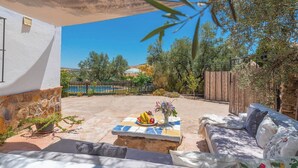 Une terrasse avec un mur en pierre, une table carrelée ornée de fruits et de fleurs, un canapé bleu et une vue sur une piscine et des arbres.