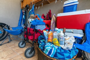 Ein Korbmöbel gefüllt mit Handtüchern und Pflegeprodukten, ein Kinderwagen, ein Fahrrad und eine rote und weiße Kühlbox auf einem Fliesenboden.