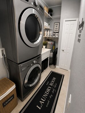 Un cuarto de lavandería con lavadoras y secadoras apiladas, un estante con productos de limpieza y una puerta con un cartel sobre lavandería.