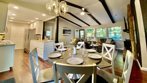 Un'ampia sala da pranzo con soffitto alto e travi a vista, una cucina adiacente allo spazio pranzo e un soggiorno con divano e ampie finestre.