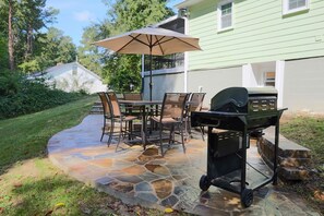 An outdoor patio with stone flooring, a table with chairs, a grill cart, and an umbrella. - Beautiful Home At Downtown Apex (Apex)
