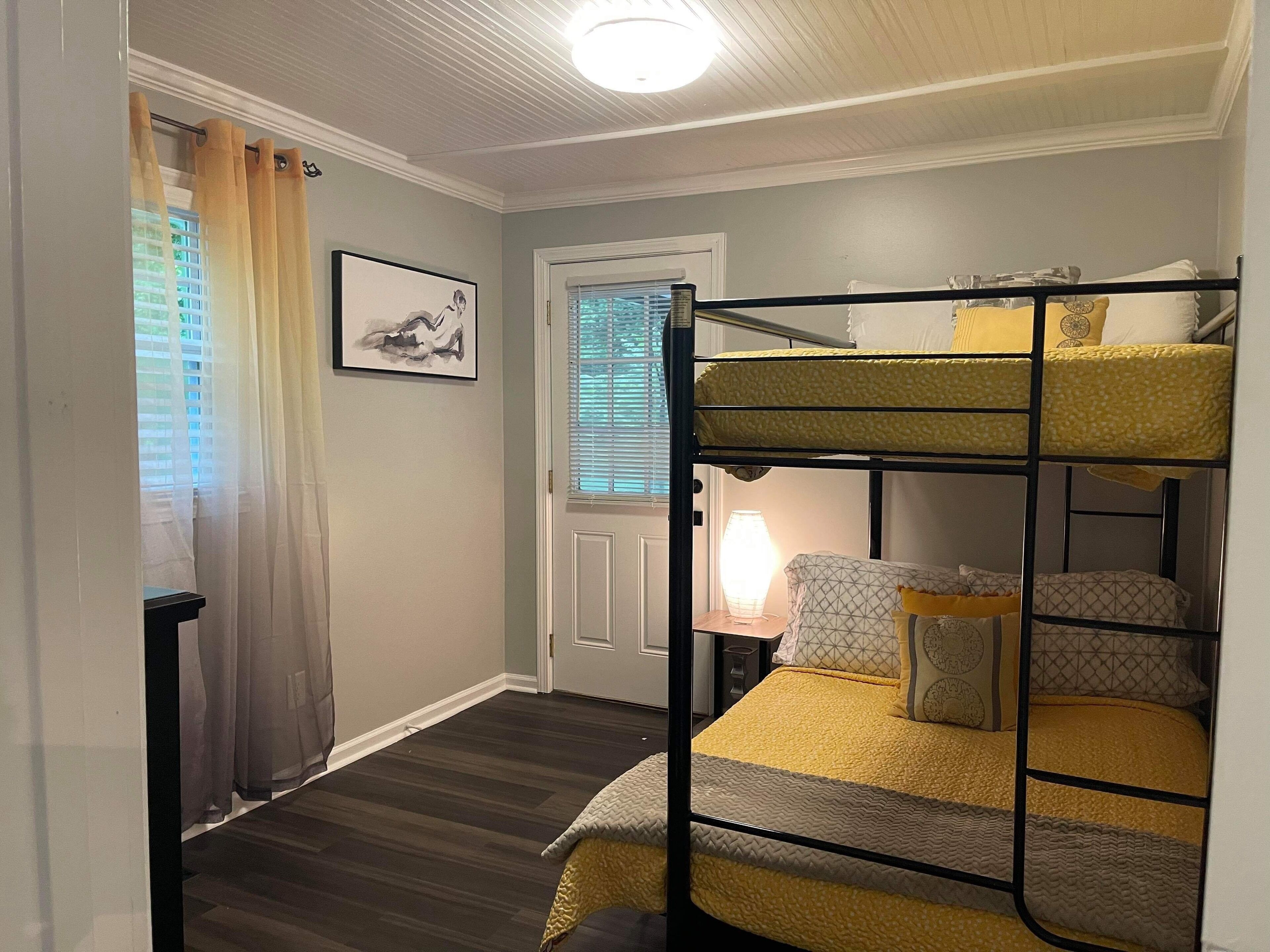 A bunk bed room with a door, a framed picture on the wall, and a lamp on a bedside table.