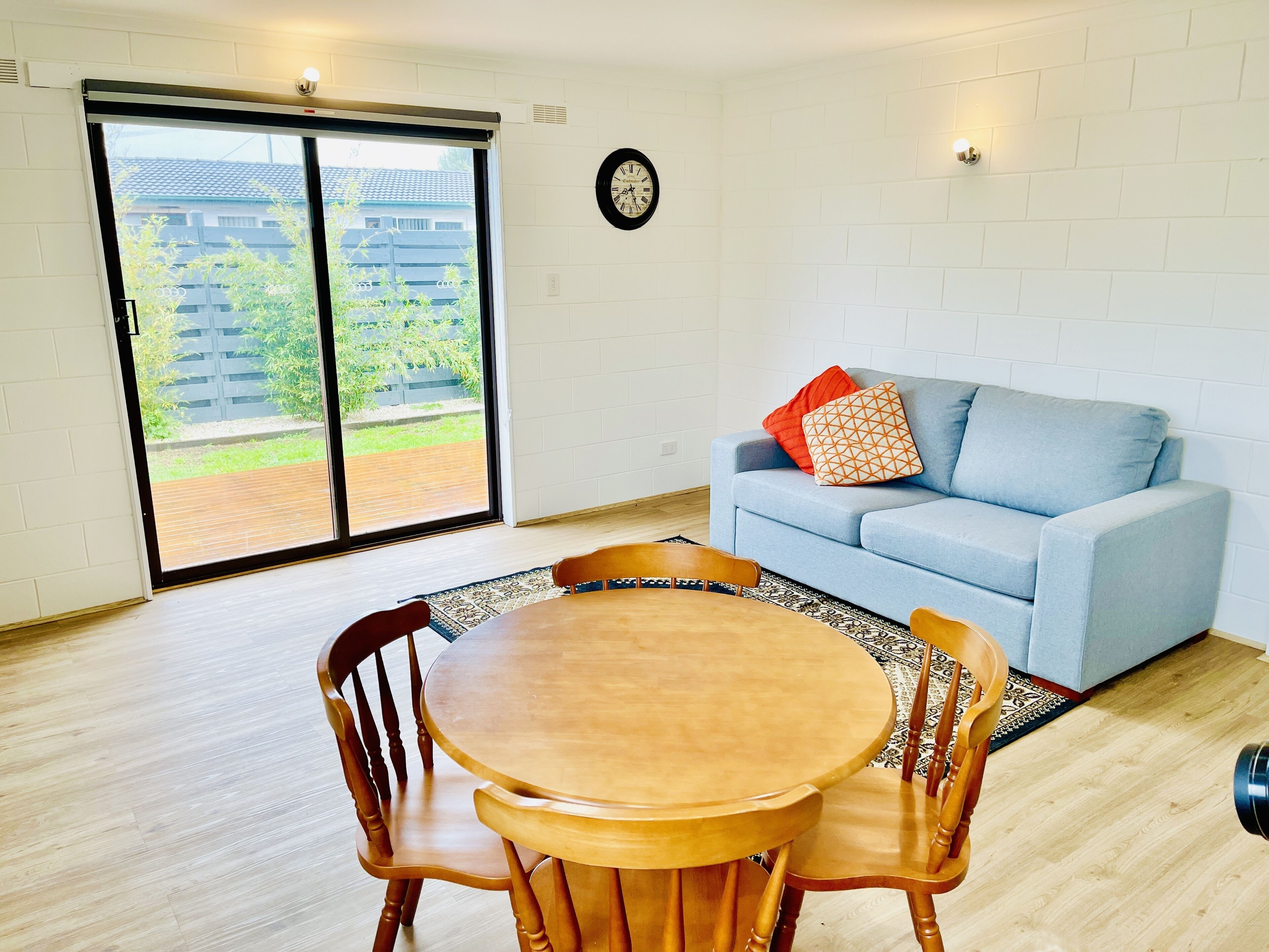 Una habitación con una mesa de comedor y sillas, un sofá con cojines, un reloj en la pared y una puerta corrediza de vidrio que da a un espacio exterior.