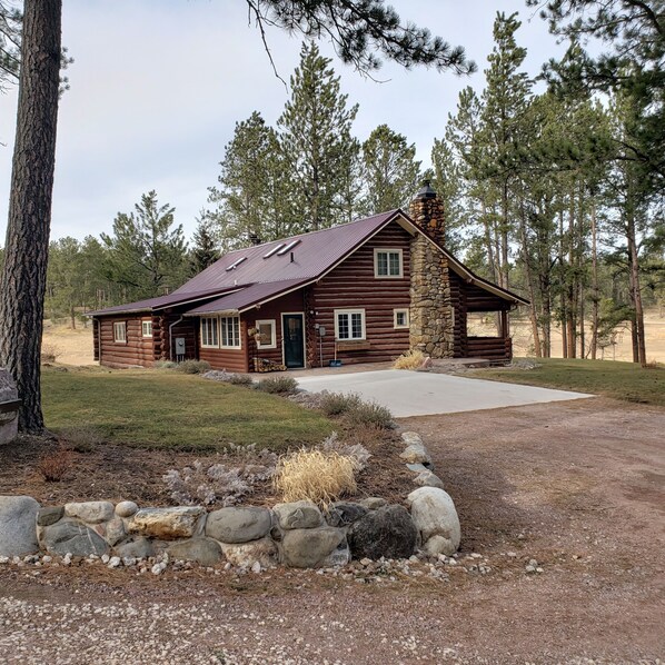 A rustic log cabin with a stone chimney, a front porch, and a gravel driveway. - Two story authentic log construction home, built in 1936 with two fireplaces. (Custer)