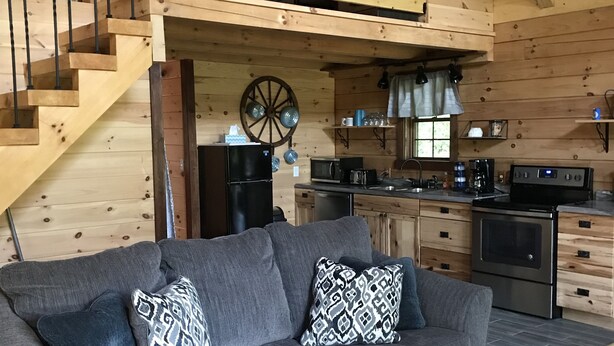 A cozy cabin interior with a grey sofa, wooden staircase, kitchen area, and a dining table.