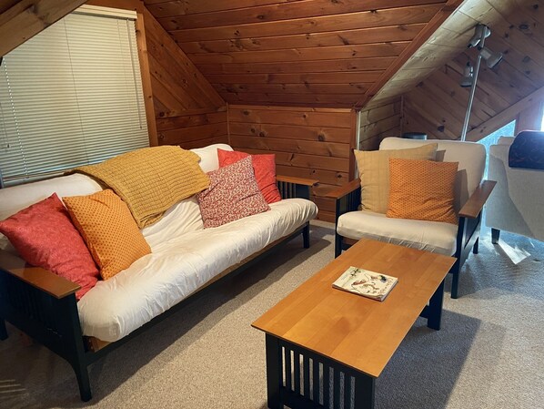 A cozy room with a wooden ceiling, a white sofa with patterned cushions, a wooden coffee table, and a white armchair.