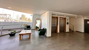 Een ruime lobby met een houten tafel, groene stoelen, een grote spiegel en een lift.