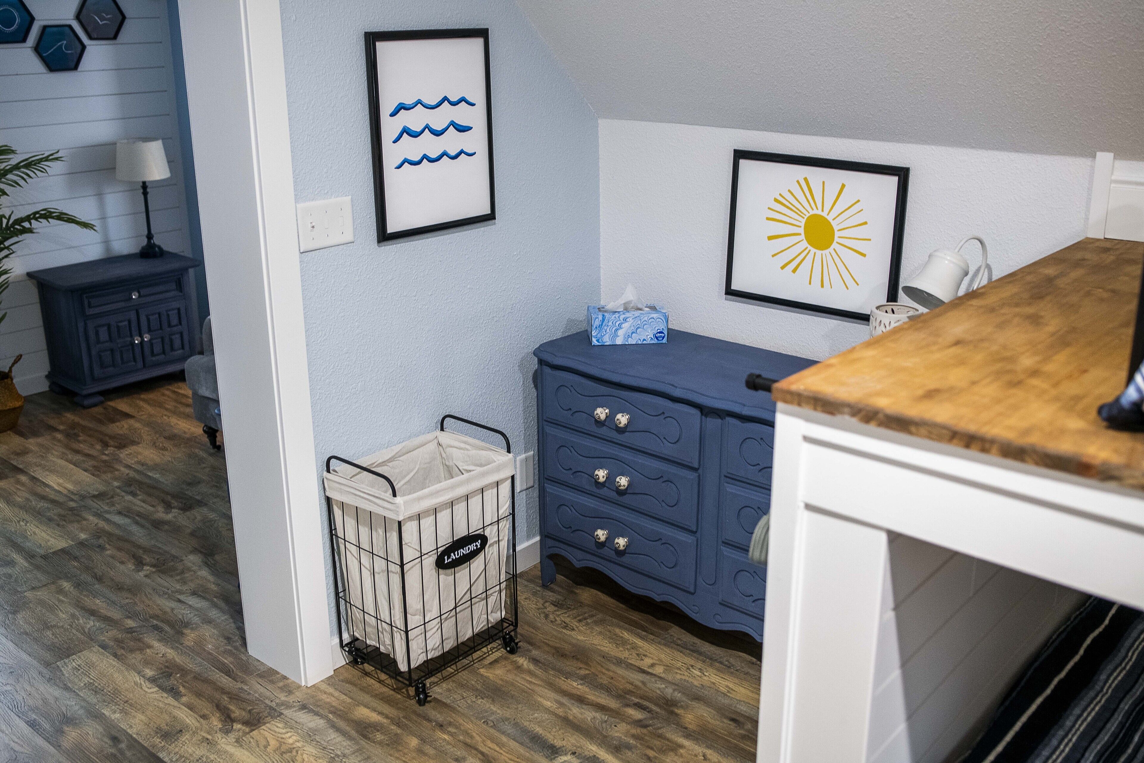 Una habitación con piso de madera, un aparador azul, un armario blanco, un cesto de ropa y un cuadro con olas y un sol.