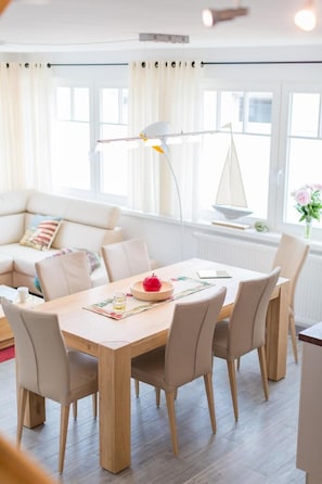 Ein modernes Wohnzimmer mit einem Holzesstisch, beigen Stühlen, einem weißen Sofa und einer Segelbootsdekoration auf der Fensterbank.