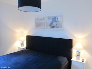 Et soveværelse med sort sengegavl, seng med blå dyne, to sengeborde med lamper og et billede hængt på væggen.