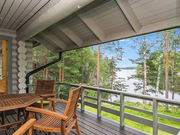 A wooden deck with a table and chairs, overlooking a lake with trees and a clear sky.