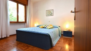 A bedroom with a large bed, a window with curtains, a bedside table with a lamp, and a painting on the wall. - Wonderful  villa for 8 people with A/C, TV, terrace and pets allowed (Lignano Sabbiadoro)