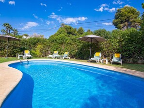 Piscina con sillones de descanso y sombrillas, rodeada de vegetación.