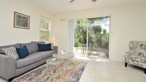 Ein Wohnzimmer mit grauem Sofa, einem Glastisch, einem gemusterten Sessel, einem Deckenventilator und einer Glasschiebetür zu einem Außenbereich.