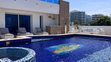 A swimming pool with lounge chairs and a hot tub, surrounded by a modern building with large windows and a stone wall.