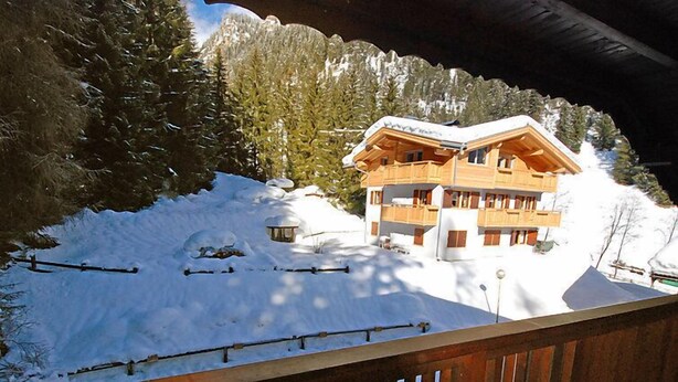 Eine Holzchalet mit mehreren Balkonen, umgeben von schneebedeckten Bäumen und einem Bergpanorama.