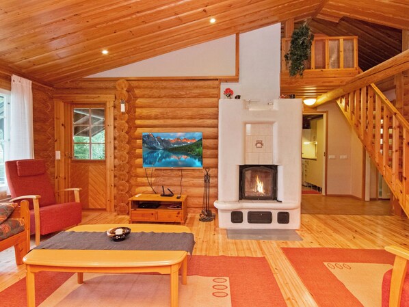 Interior de una cabaña de troncos con chimenea, un televisor montado en la pared y una zona de estar acogedora con una butaca roja y un banco de madera.