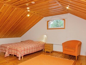 Un pequeño y acogedor dormitorio en el ático, con techo de madera inclinado, una cama individual, una mesita de noche, una silla y una ventana que proporciona luz natural.