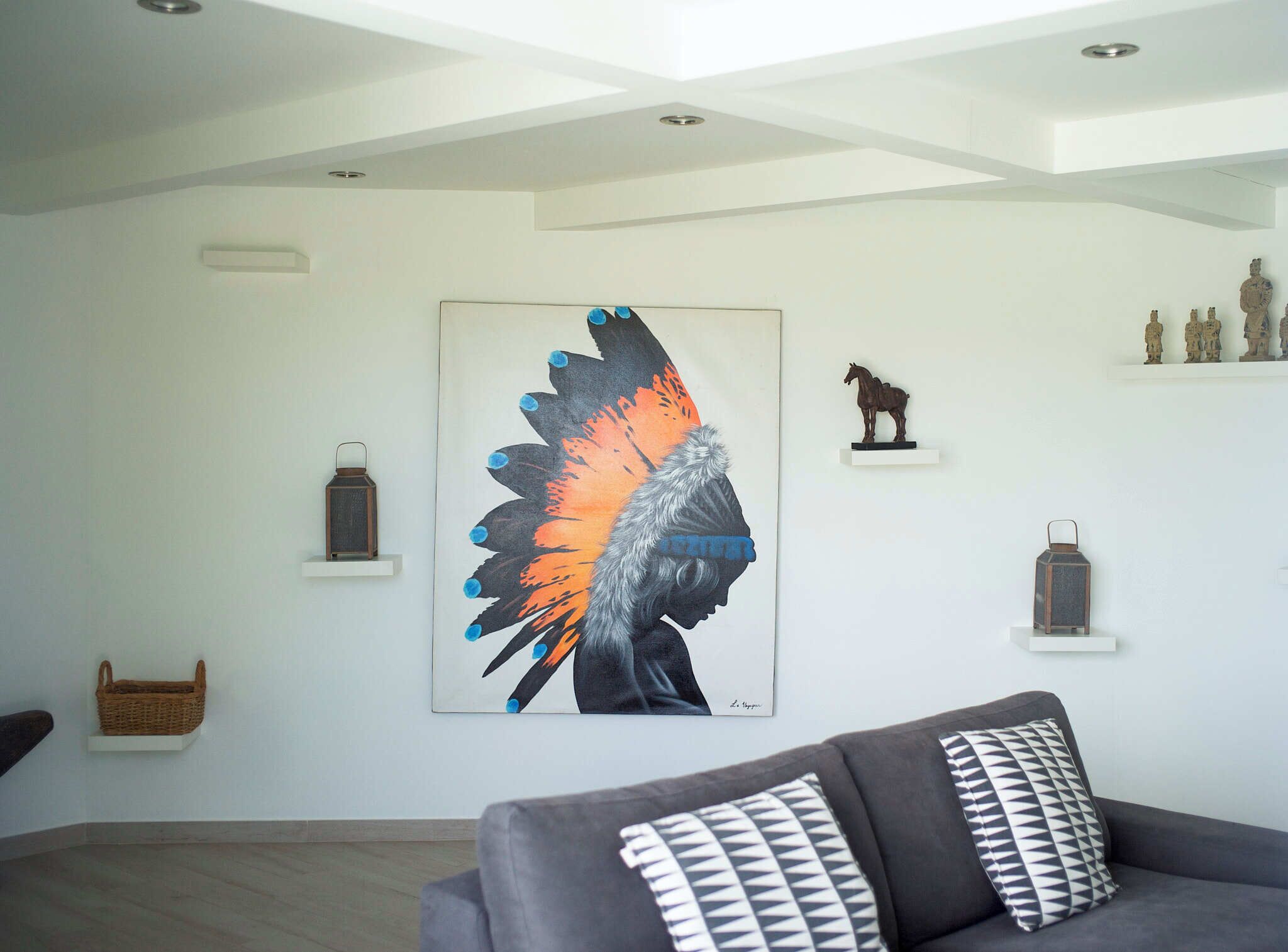 A modern living room with a large abstract painting, a grey sofa with patterned cushions, and a small shelf with decorative items.