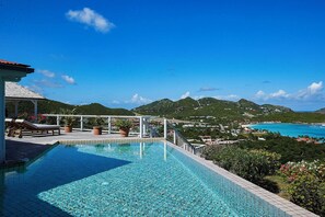 Ein Pool mit klarem, blauem Wasser, umgeben von einer weißen Uferreling und Topfpflanzen. Im Hintergrund erheben sich grüne Hügel und eine Küstenlandschaft mit Gebäuden.