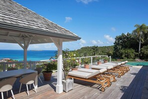 Eine Holzterrasse mit Liegestühlen und einem Tisch, mit Blick auf einen Pool und den Ozean.