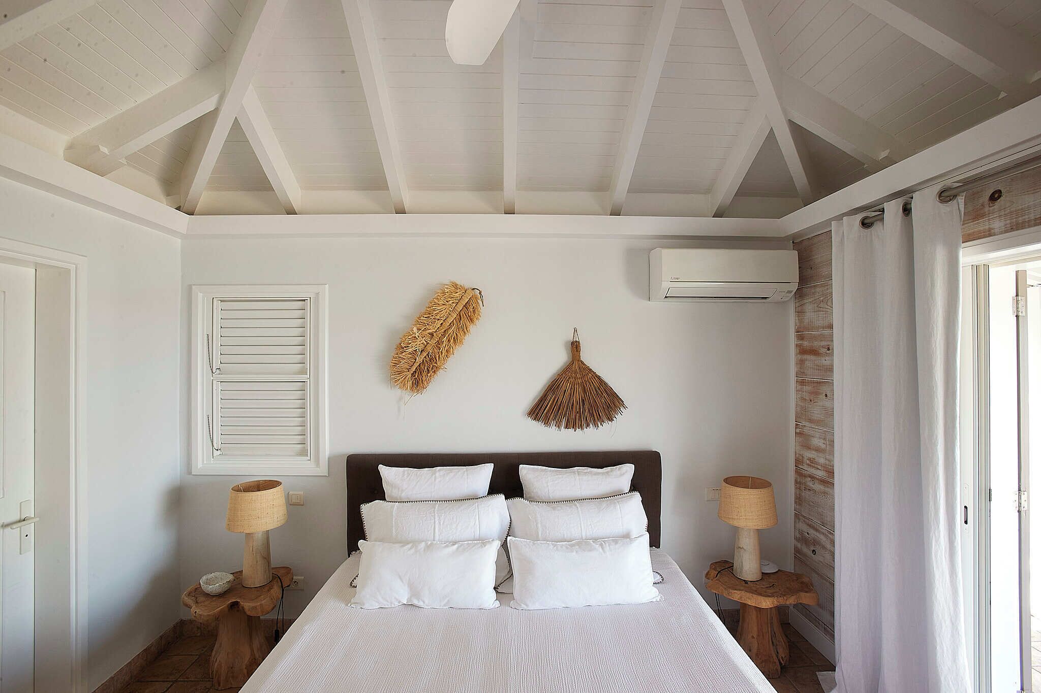 A bedroom with a large bed, white walls, wooden beams on the ceiling, a wall-mounted air conditioner, and a window with curtains.