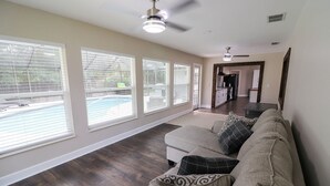 A living room with a beige sofa, patterned cushions, and large windows overlooking a backyard with a pool. - Sandy Toes Paradise (Palm Coast)