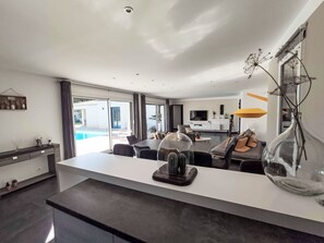 A modern living room with a large glass door leading to a pool, a dining area with a glass table, and a kitchen with a TV and white cabinets. - Cedelen by Interhome (Plan de la Tour)