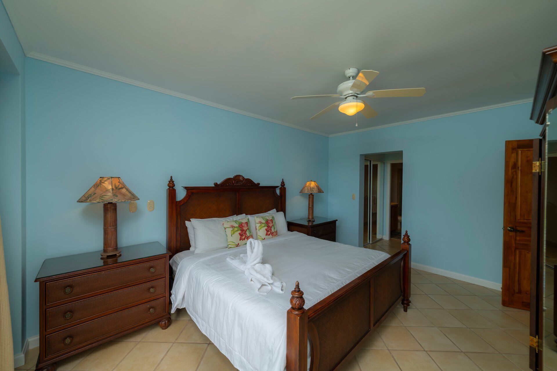 A bedroom with a wooden bed, two bedside tables with lamps, a ceiling fan, and a door.