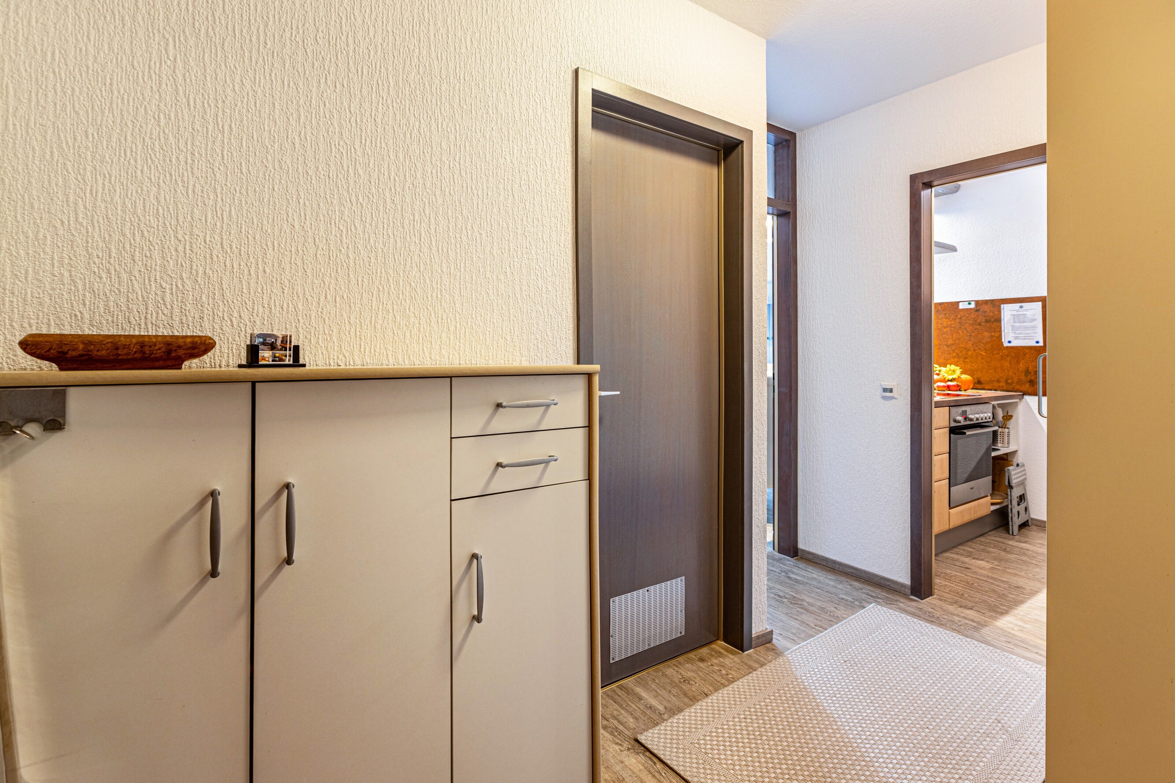 Un couloir avec une armoire blanche, un bol en bois posé sur le dessus, et une porte donnant sur un espace cuisine.